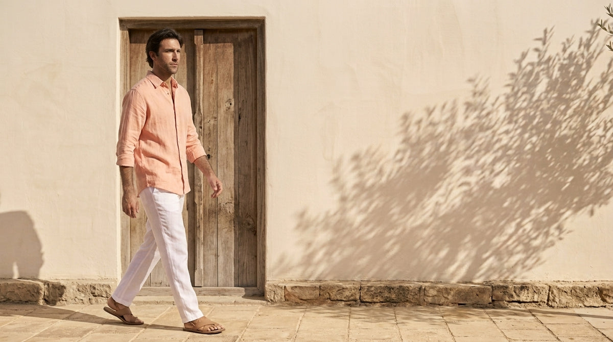 Man walking in a pink hemp shirt and white trousers past a wooden door in a sunlit courtyard
